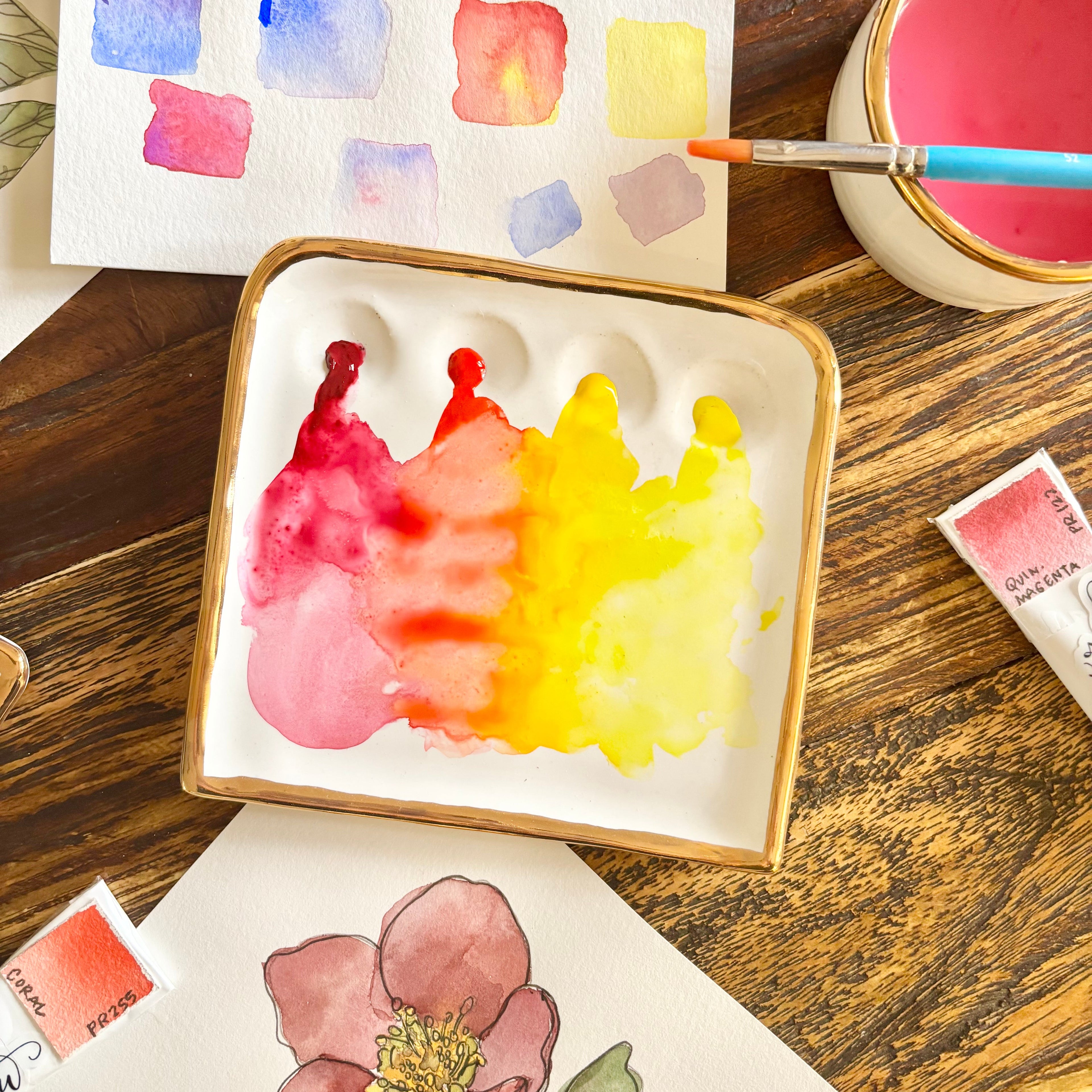 An overhead view of "The Kris Palette". This palette is a small white porcelain painter's palette with four paint wells, a spacious central mixing area, and Eleni's signature 22k gold edge. The palette is shown in use with vibrant paints being mixed in the central mixing area from the paint wells around the center.