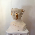 A front 45º angle view of a ceramic bust titled "She Who Sheds". The bust features a 22k gold trimmed white flower behind the ear and gold tears running down the face from the eyes. Additional gold stroke accents provide further details across the piece.