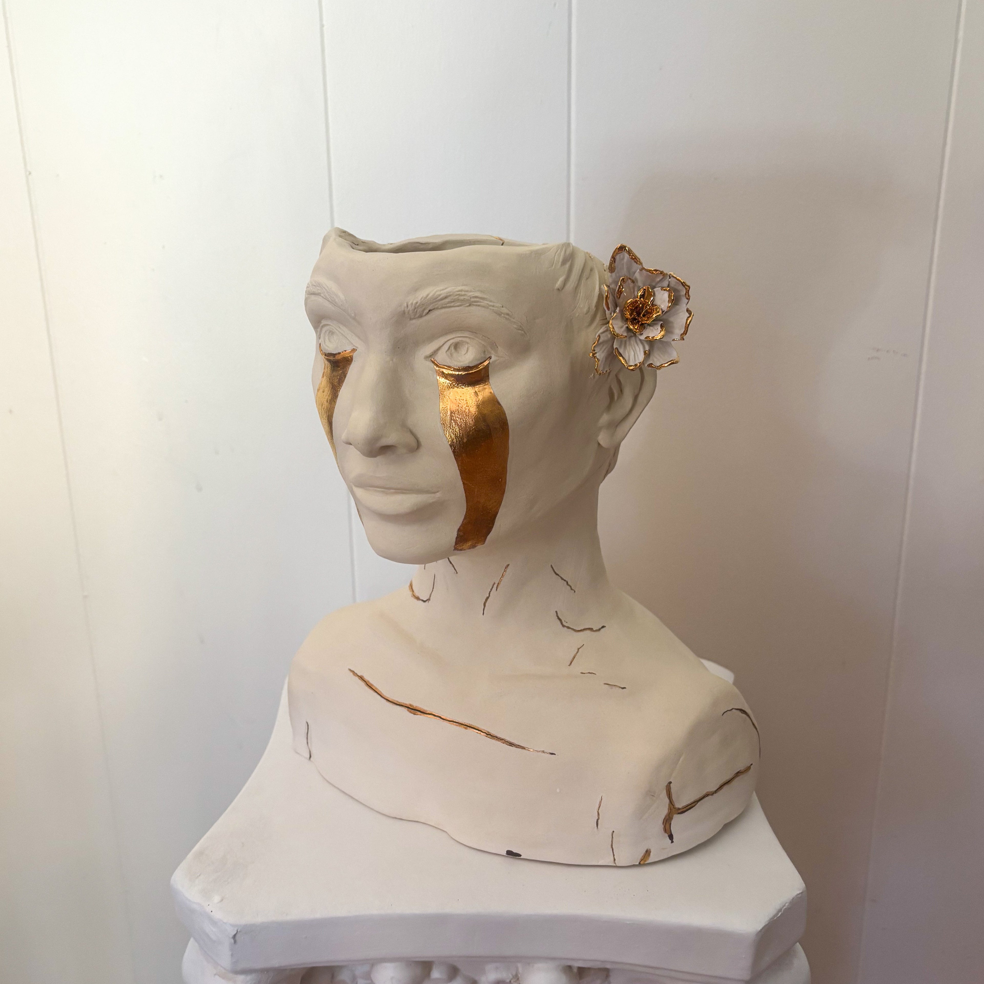 A front 45º angle view of a ceramic bust titled "She Who Sheds". The bust features a 22k gold trimmed white flower behind the ear and gold tears running down the face from the eyes. Additional gold stroke accents provide further details across the piece.