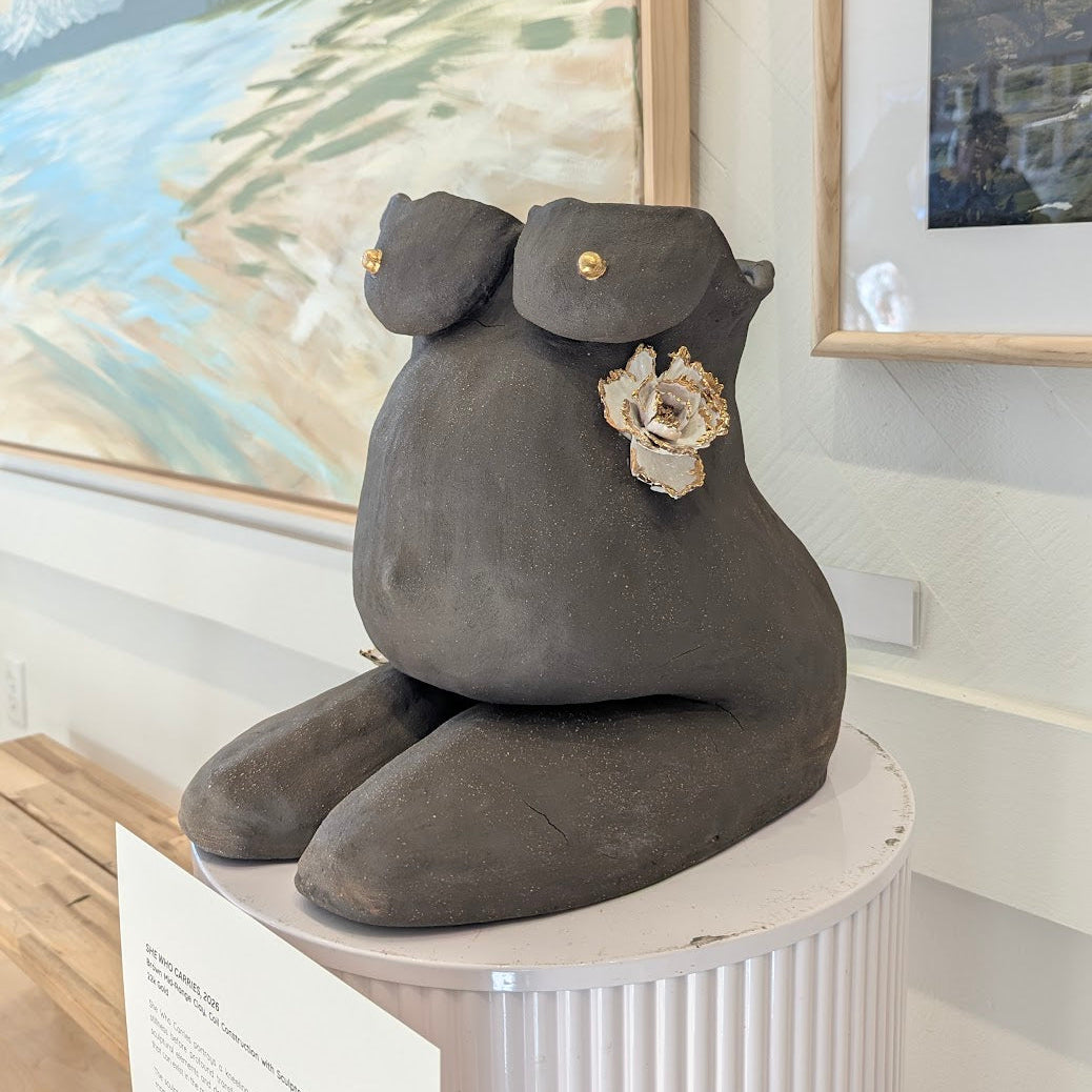 A 45º-angle view of the clay sculpture "She Who Carries" by Eleni. The grey piece is adorned with 22k gold trimmed white flowers. The sculpture is in a kneeling position.
