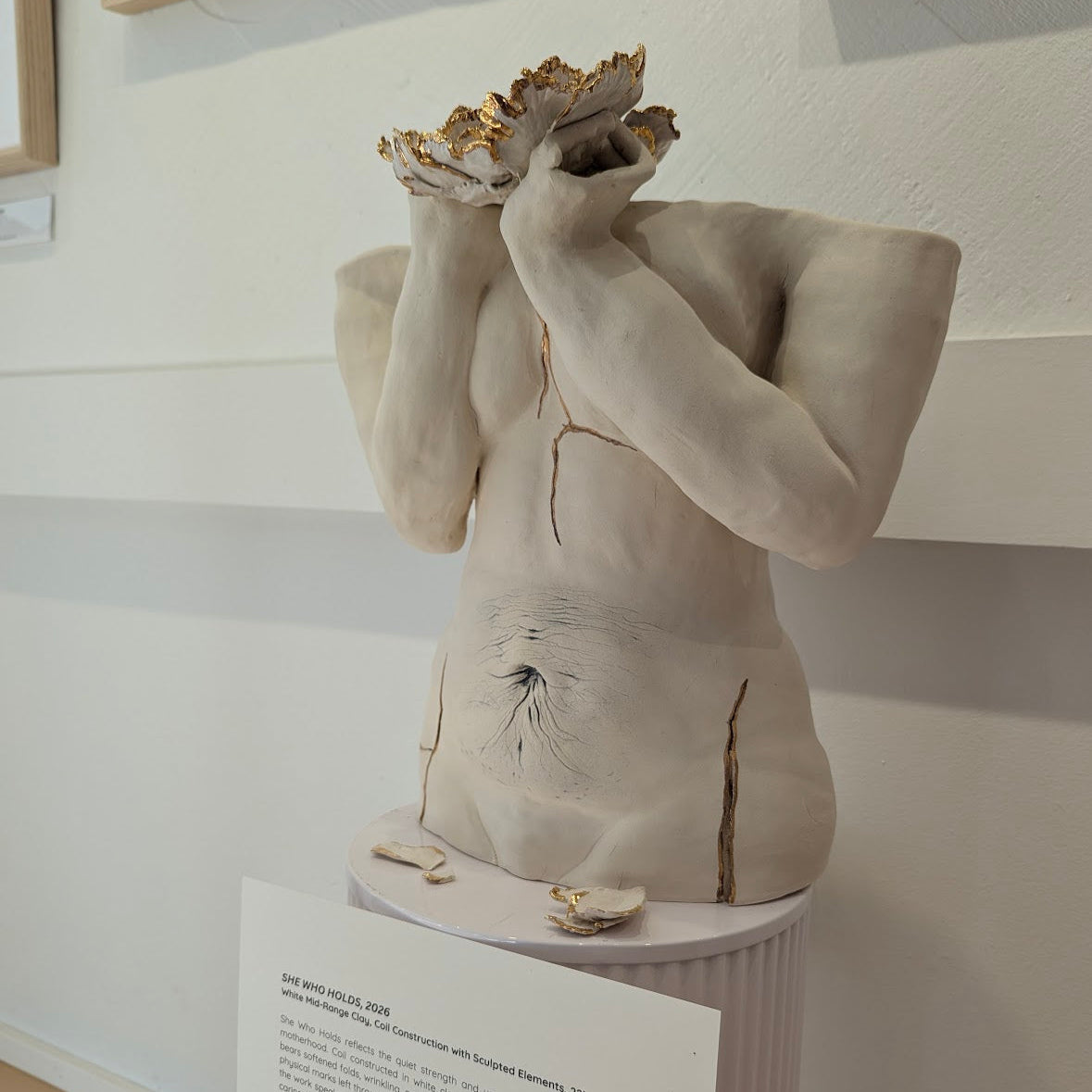 A front off-center angle view of a ceramic sculpture titled "She Who Holds". The sculpture features floral elements, in addition to softened folds, wrinkling, and stretched surfaces that reference the lasting physical marks left upon the body through carrying and nurturing a child. Throughout the piece are 22k gold stroke details, signature to Eleni's artistic style.