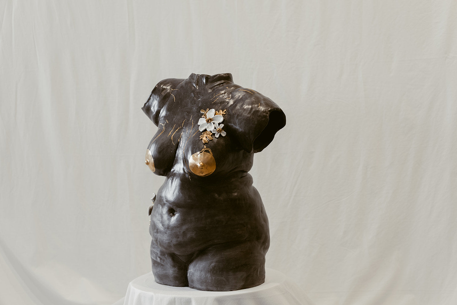 A 45º front view of a ceramic bust composed of mixed black mid-range clay titled "She Who Feels" by Eleni. The sculpture is adorned with hand-sculpted white flowers that and accents of 22k gold.
