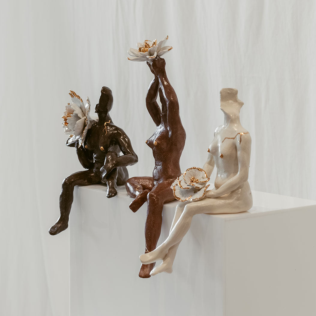 Three figurative sculptures displayed on a white pedestal against a white draped backdrop. From left to right, the female figures are dark, terracotta, and white ceramic.