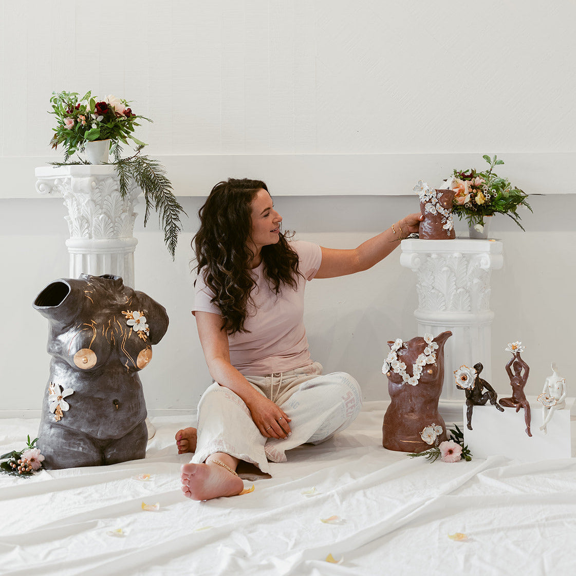 Eleni, barefoot on the studio floor, surrounded by sculptures of various sizes and in various colors. There are also plants in the composition, adding organic color.