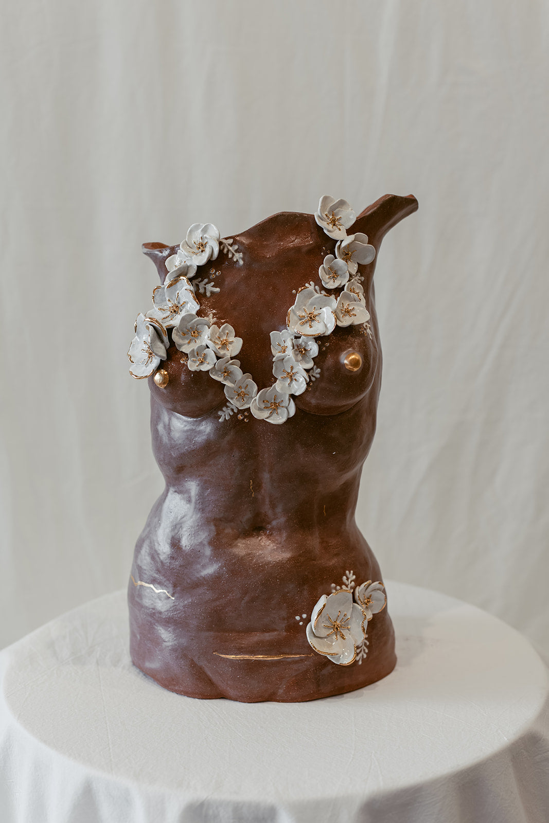 Frontal view view of a ceramic bust called "She Who Remains" by Eleni. The sculpture features 22k gold trimmed white sculpted floral elements across the chest and lower body. The piece bears subtle cracking and the presence of a c-section scar, accented by 22k gold strokes.