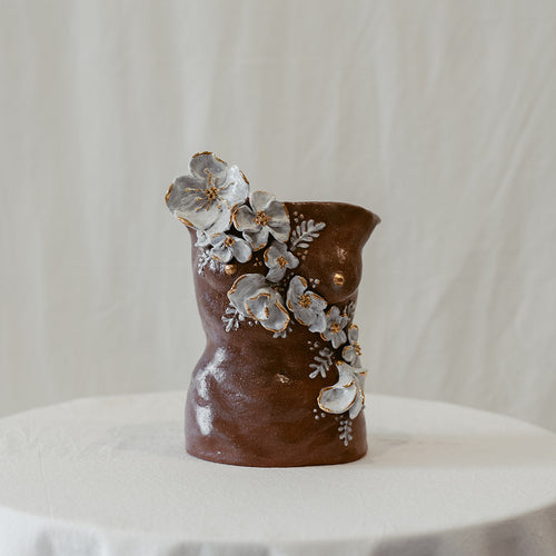 A straight-on view of a ceramic bust titled "She Who Waits" by Eleni. The sculpture features cascading hand-sculpted white flowers that move across the form with accents of 22k gold.