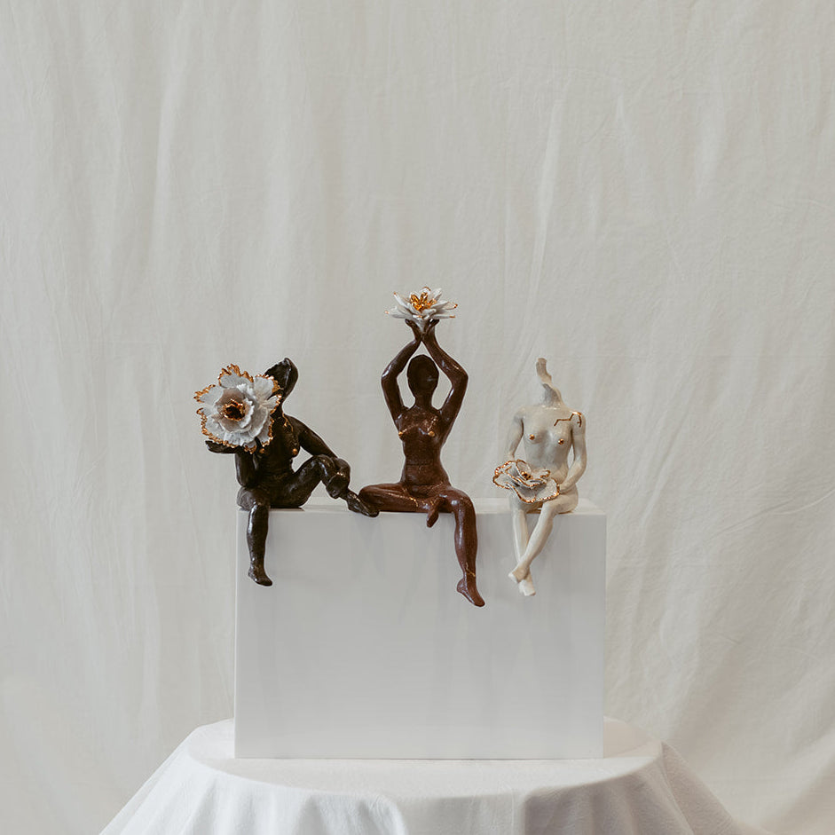 A straight-on view of "She Who Gathers" by Eleni. The piece features three individual figures formed from deep brown clay, terracotta, and white porcelain  (left to right). Each figure has 22k gold accents, including a gold trimmed floral element.