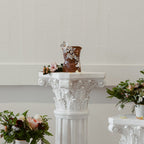 A distant straight-on view of "She Who Waits". The work is displayed on a floral column pedestal with flowers.