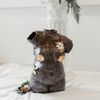 A straight-on view of a ceramic bust composed of mixed black mid-range clay titled "She Who Feels" by Eleni. The sculpture is adorned with hand-sculpted white flowers that and accents of 22k gold.