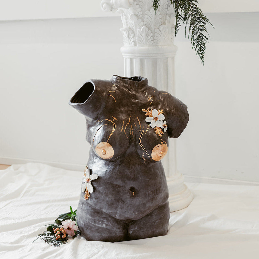 A straight-on view of a ceramic bust composed of mixed black mid-range clay titled "She Who Feels" by Eleni. The sculpture is adorned with hand-sculpted white flowers that and accents of 22k gold.