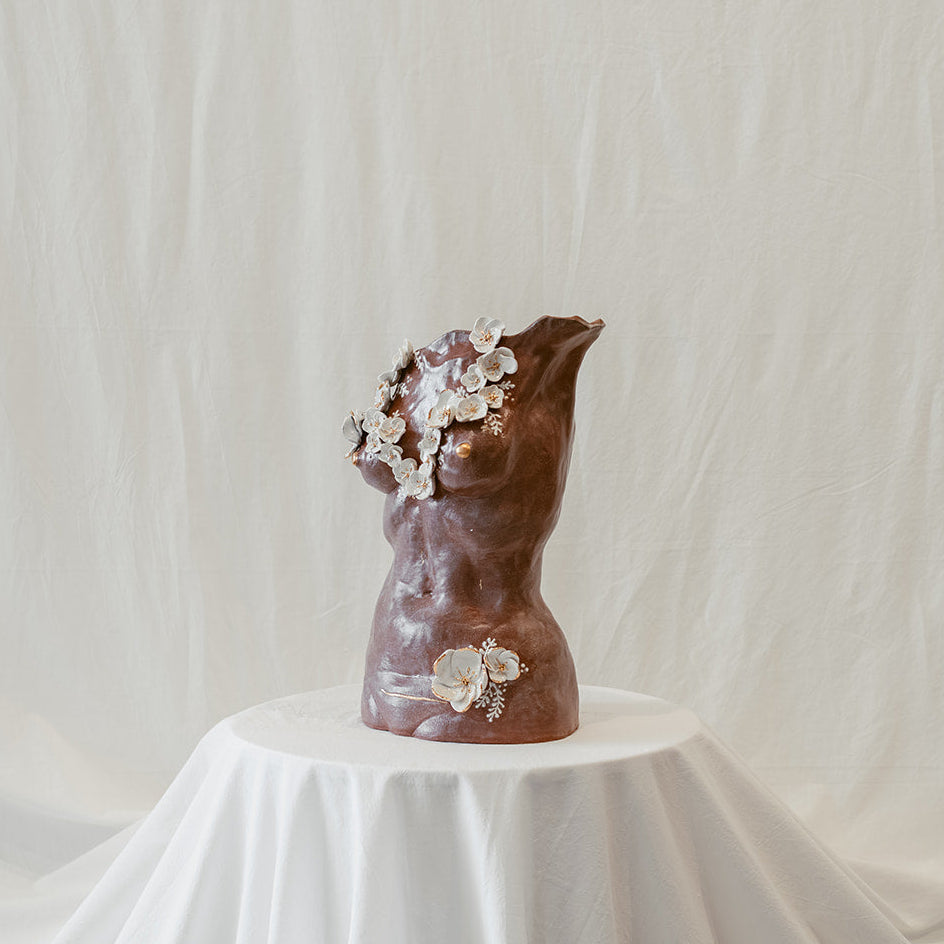 A 45º angle view of a ceramic bust called "She Who Remains" by Eleni. The sculpture features 22k gold trimmed white sculpted floral elements across the chest and lower body. The piece bears subtle cracking and the presence of a c-section scar, accented by 22k gold strokes.
