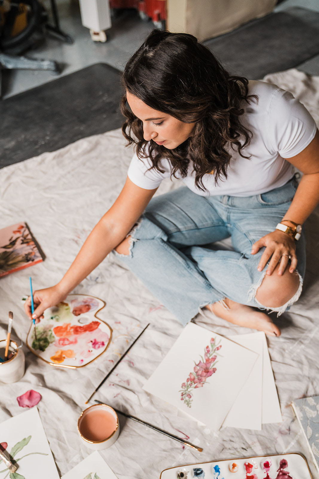 Eleni with painter’s palette she made. The palette is white ceramic with a gilded edge. There is paint, and she is using it to paint flowers on paper. There is also a ceramic cup for paint water.