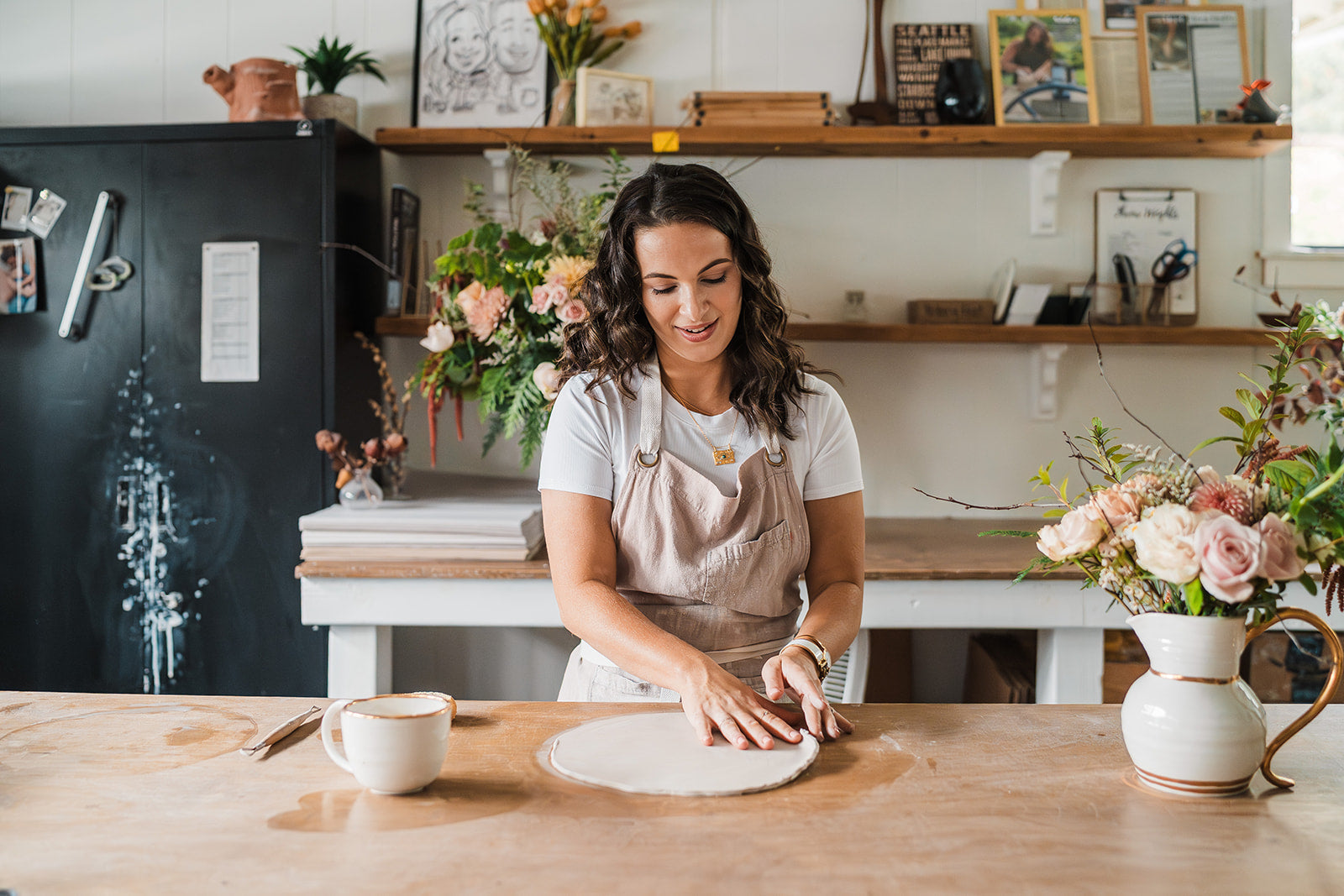 Eleni wearing an apron, rolling out clay on table in a decorated studio space with shelves, framed art, and flower arrangements.