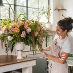 Eleni stands beside a large floral arrangement in a white pedestal vase on a wooden worktable inside a bright, sunlit craft studio.