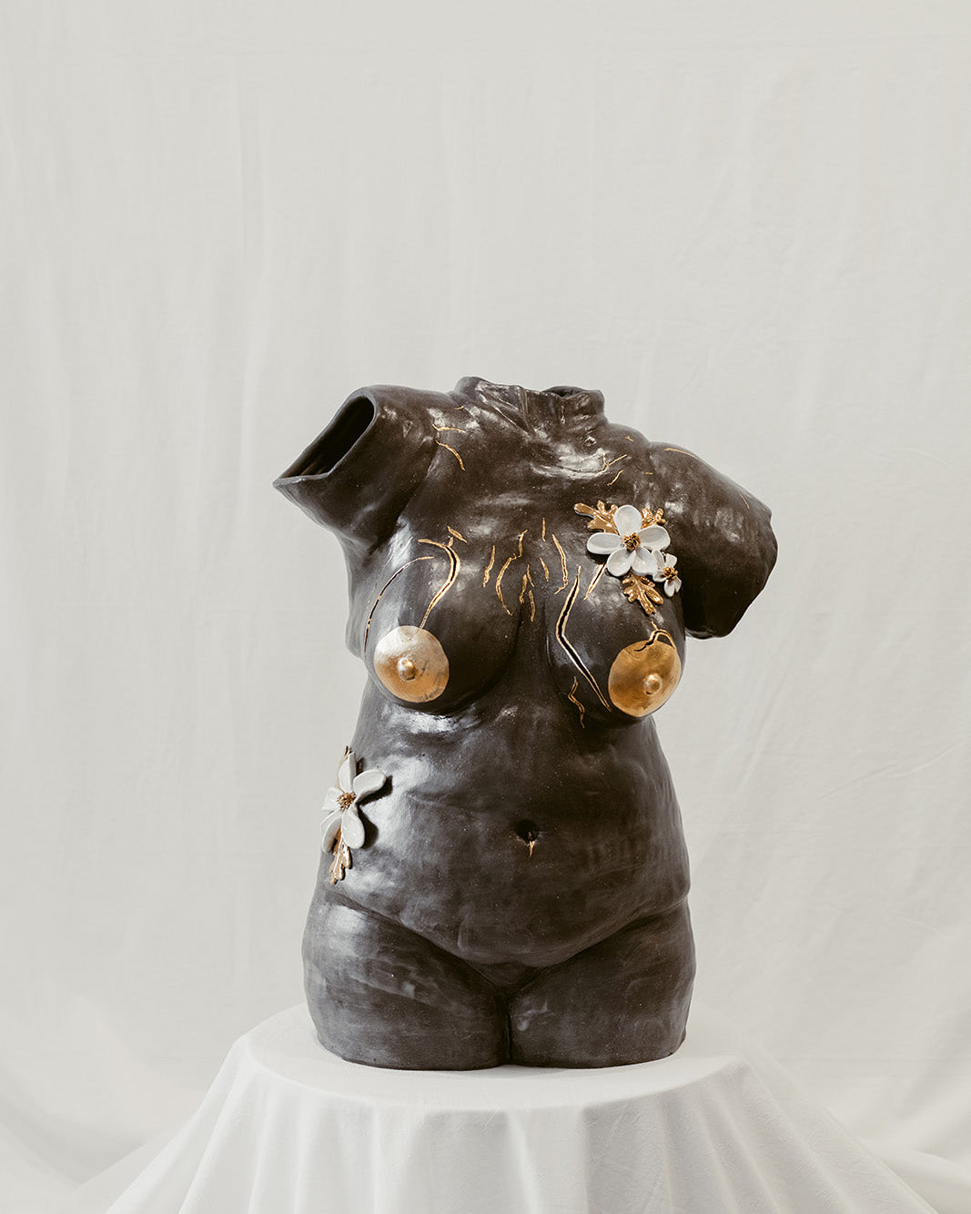 A straight-on view of a ceramic bust composed of mixed black mid-range clay titled "She Who Feels" by Eleni. The sculpture is adorned with hand-sculpted white flowers that and accents of 22k gold.