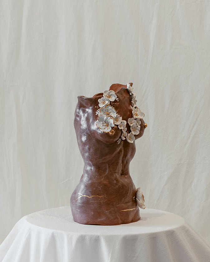 A 45º angle view of a ceramic bust called "She Who Remains" by Eleni. The sculpture features 22k gold trimmed white sculpted floral elements across the chest and lower body. The piece bears subtle cracking and the presence of a c-section scar, accented by 22k gold strokes.