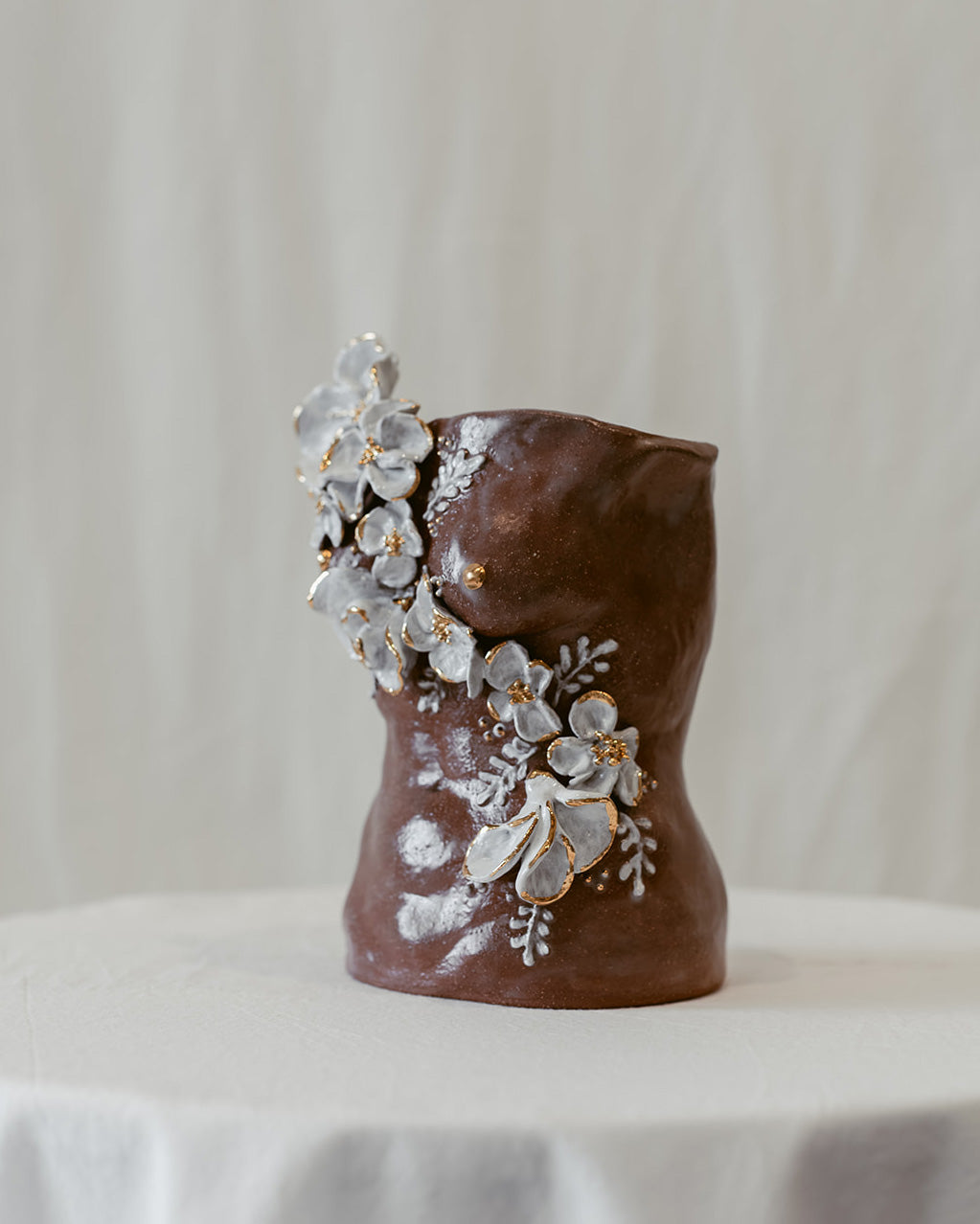 A 45º side view of a ceramic bust titled "She Who Waits" by Eleni. The sculpture features cascading hand-sculpted white flowers that move across the form with accents of 22k gold.