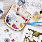 An overhead view of Eleni's "Vienna Painter Travel Palette". The palette is a white porcelain painter's palette with Eleni's signature 22k gold edge. The palette features five paint wells and a large mixing surface, displayed alongside paint brushes and artistic illustrations.