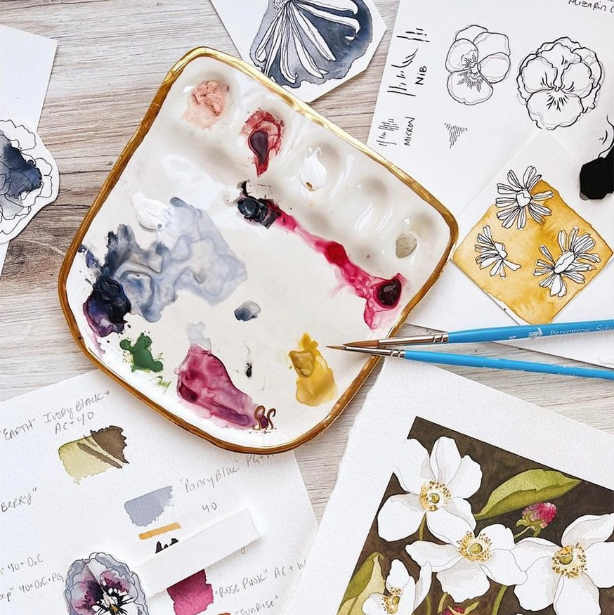 An overhead view of Eleni's "Vienna Painter Travel Palette". The palette is a white porcelain painter's palette with Eleni's signature 22k gold edge. The palette features five paint wells and a large mixing surface, displayed alongside paint brushes and artistic illustrations.