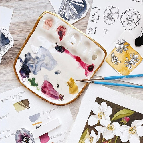 An overhead view of Eleni's "Vienna Painter Travel Palette". The palette is a white porcelain painter's palette with Eleni's signature 22k gold edge. The palette features five paint wells and a large mixing surface, displayed alongside paint brushes and artistic illustrations.
